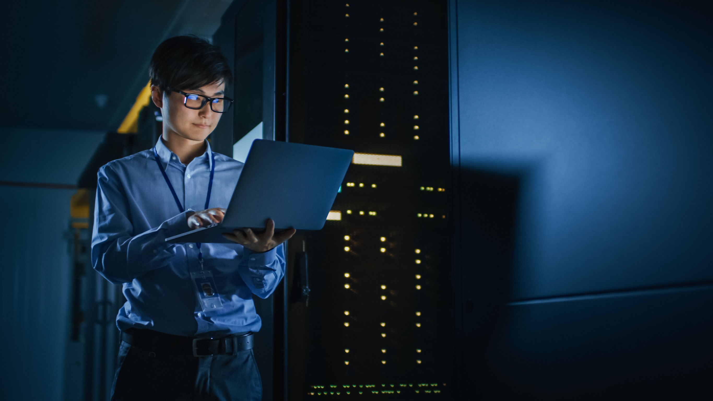 In Dark Data Center: Male IT Specialist Stands Beside the Row of Operational Server Racks, Uses Laptop for Maintenance. Concept for Cloud Computing, Artificial Intelligence, Supercomputer